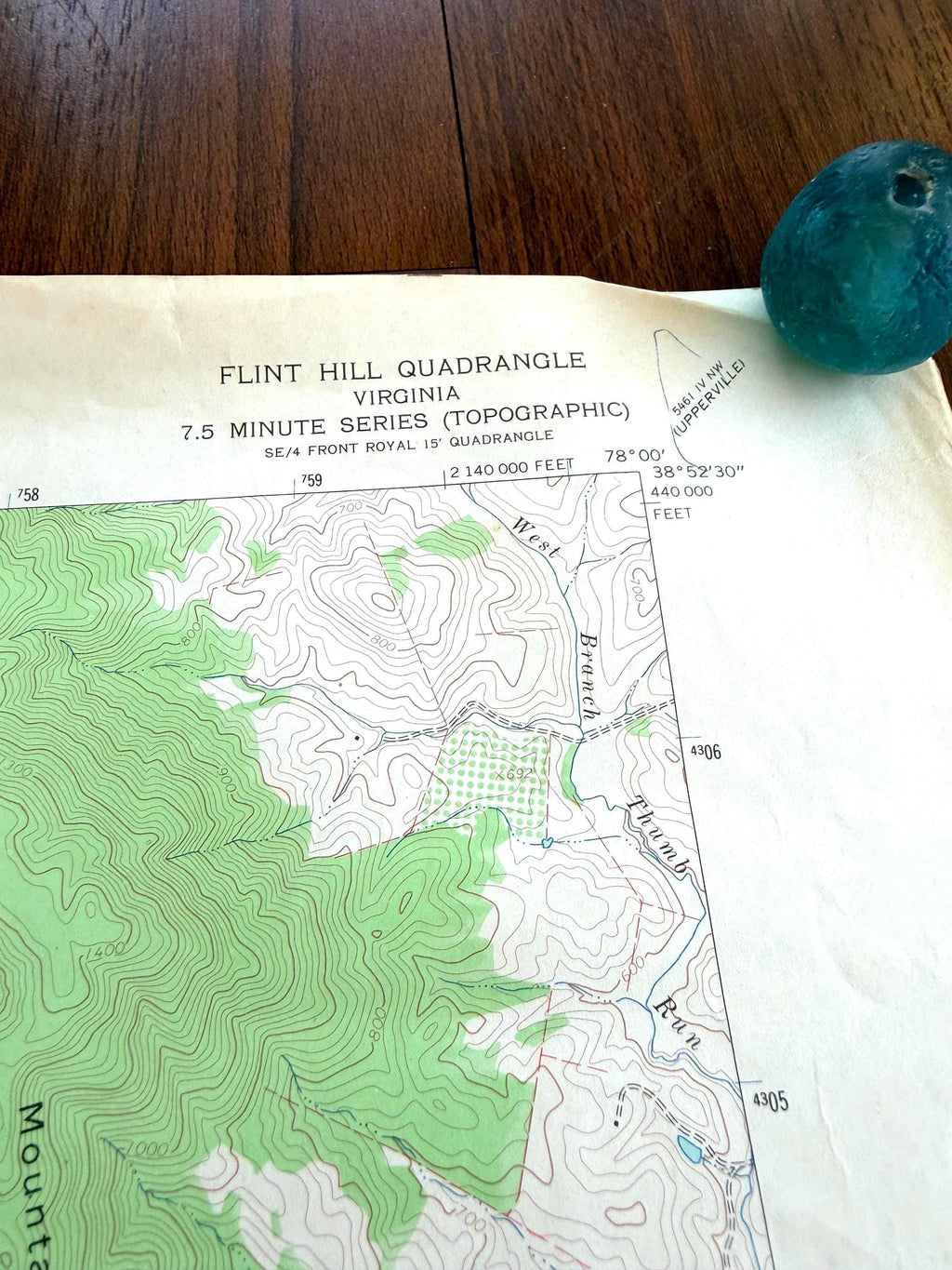 May include: A paper topographic map titled Flint Hill Quadrangle Virginia 7.5 Minute Series with green contour lines and elevation markings, positioned next to a small, round, translucent teal glass object with a hole in the center.