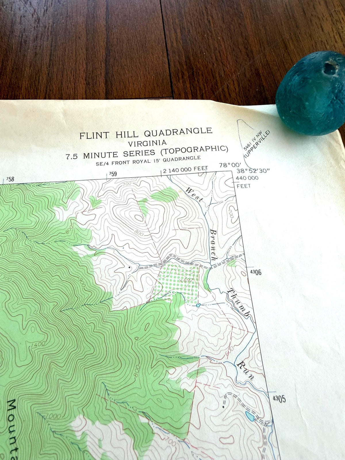 May include: A paper topographic map titled Flint Hill Quadrangle Virginia 7.5 Minute Series with green contour lines and elevation markings, positioned next to a small, round, translucent teal glass object with a hole in the center.