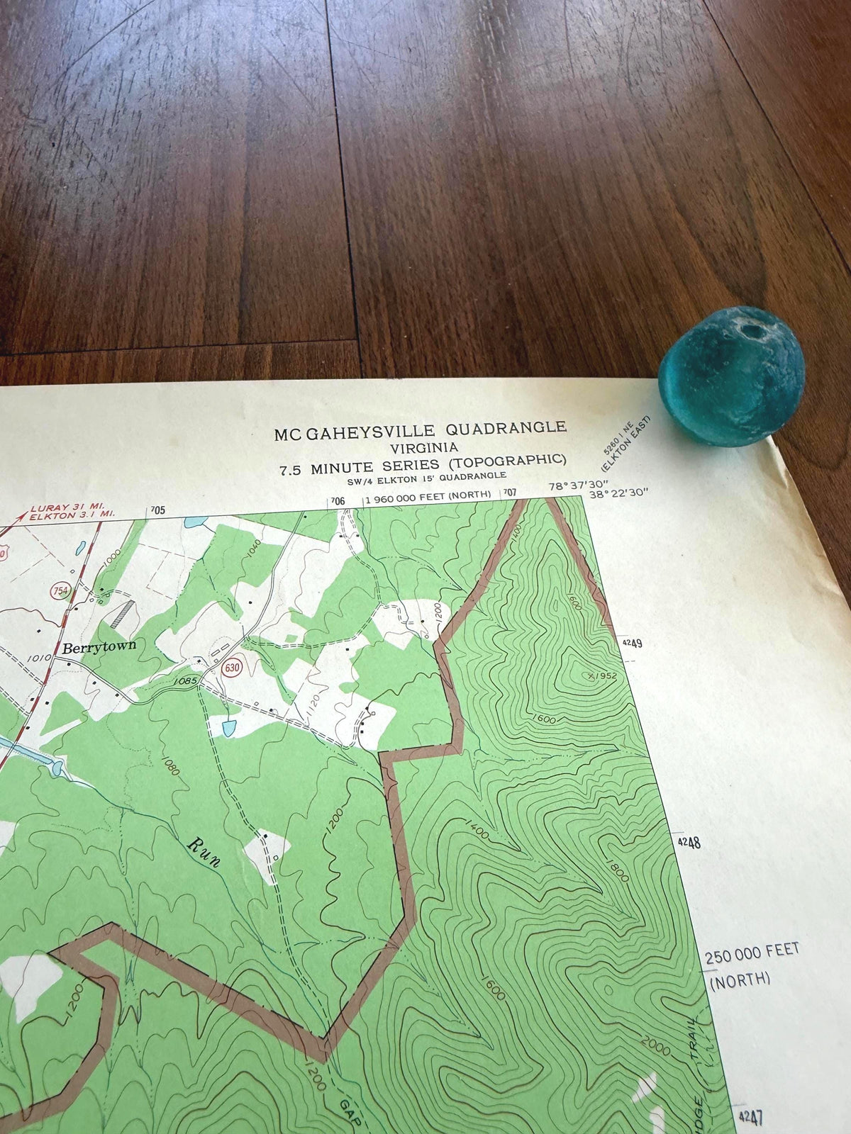May include: A topographic map of the McGaheysville Quadrangle in Virginia, 7.5 minute series, featuring green terrain contours and black text, accompanied by a small, round, translucent teal glass marble resting on the upper right corner.