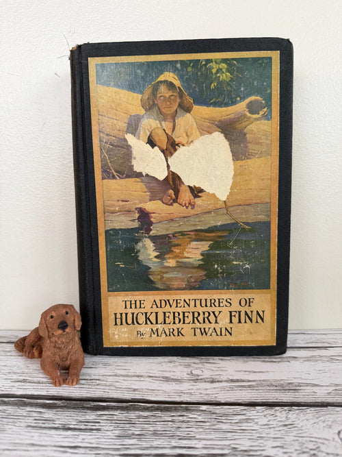 May include: An antique copy of "The Adventures of Huckleberry Finn" by Mark Twain, featuring a boy on a log illustration. A small brown dog figurine rests in front of the book on a white surface.