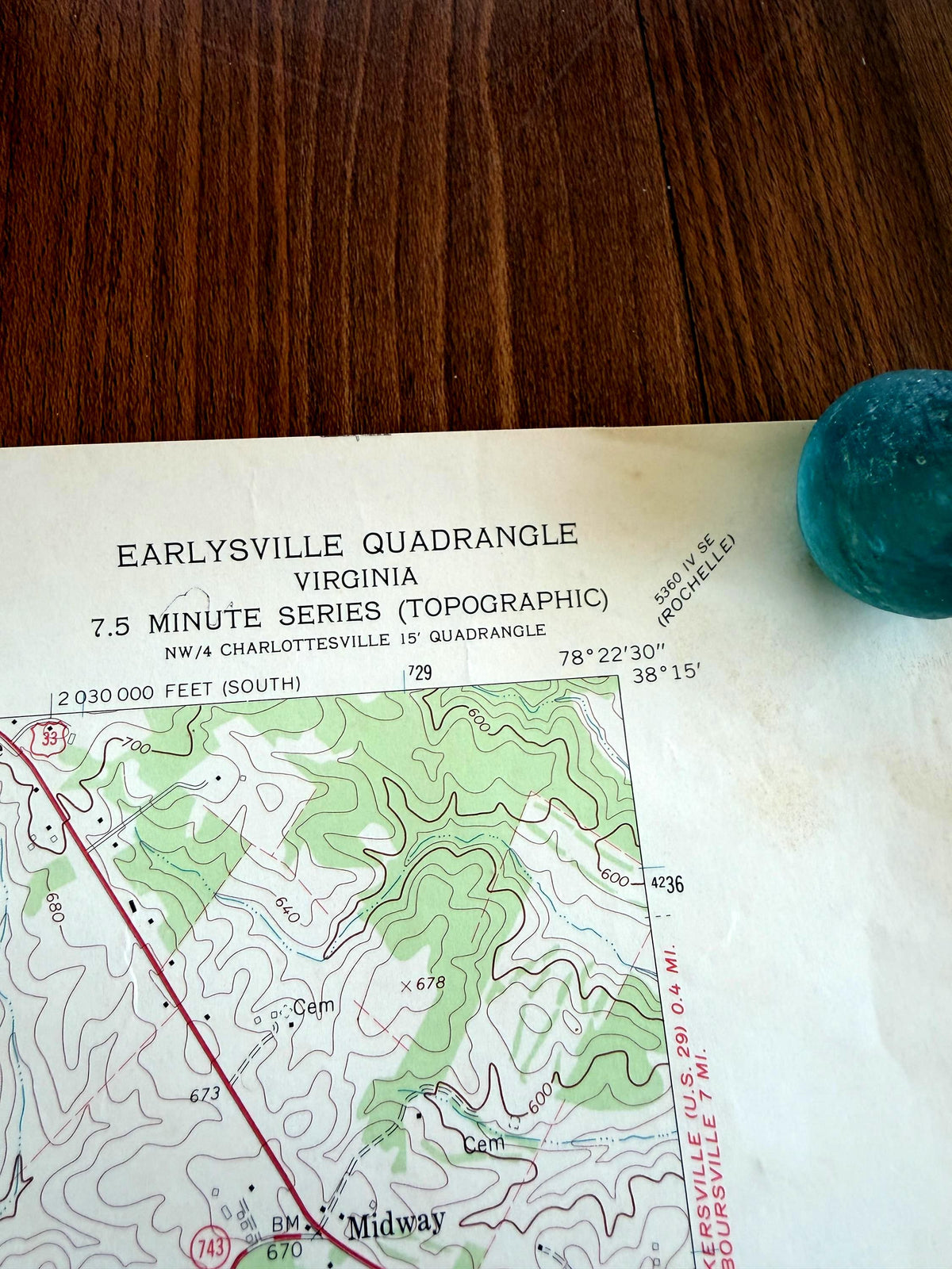 May include: A paper topographic map of the Earlysville Quadrangle in Virginia, 7.5 Minute Series, featuring contour lines, green shaded areas, and text labels. A small, round, textured teal object is positioned on the upper right corner of the map.