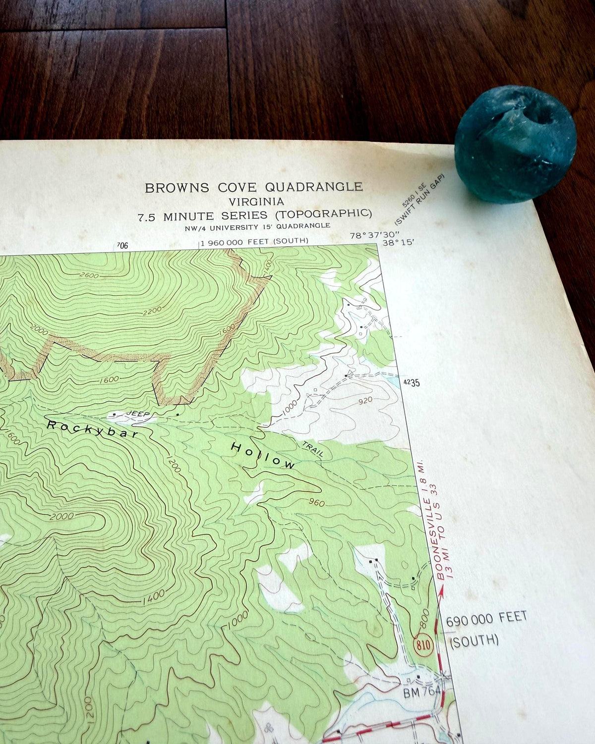 May include: A topographic map titled Browns Cove Quadrangle Virginia 7.5 Minute Series with green contour lines and text, accompanied by a small, round, teal-colored glass marble resting on the upper right corner of the paper document.