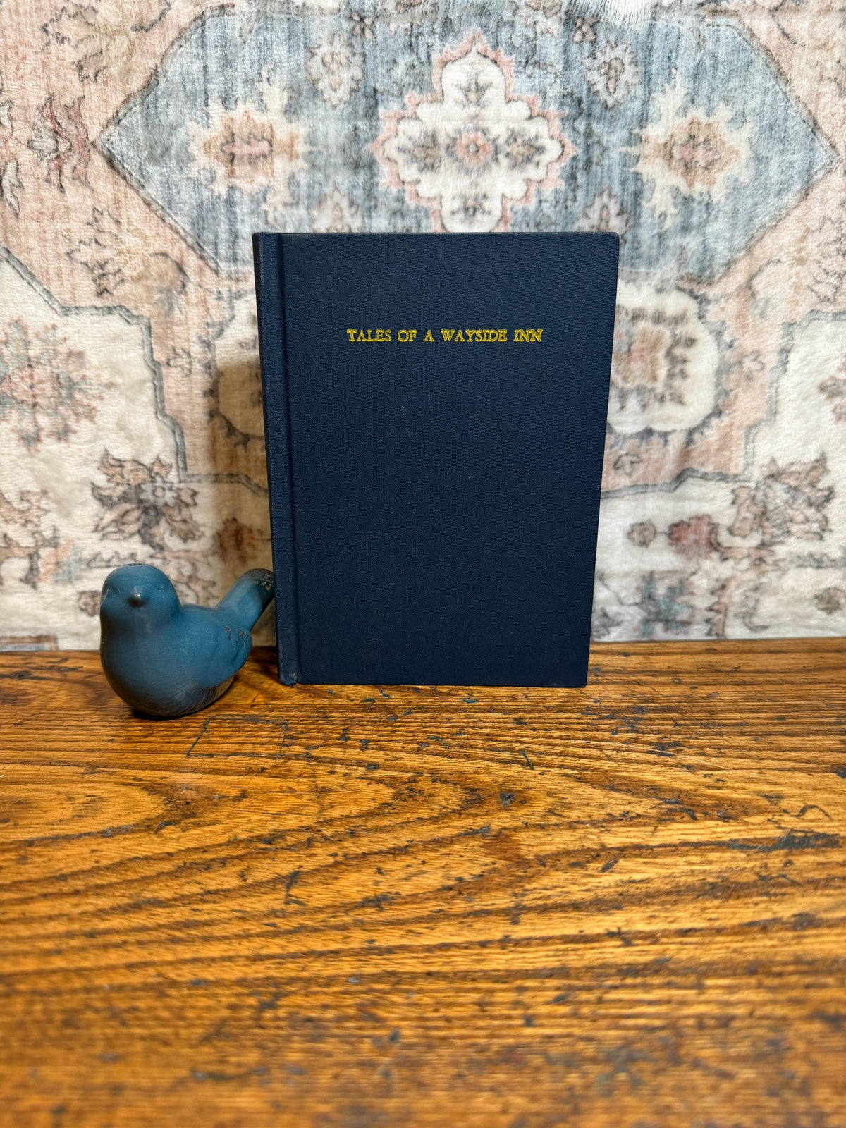 May include: A navy blue book titled "TALES OF A WAYSIDE INN" with gold lettering, next to a small blue bird figurine. The book and bird are on a wooden surface with a patterned rug in the background.