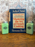 May include: A still life featuring a vintage copy of "Mastering the Art of French Cooking" by Julia Child and Simone Beck, flanked by two green glass salt and pepper shakers. The book is blue with orange text. The salt and pepper shakers have silver lids.