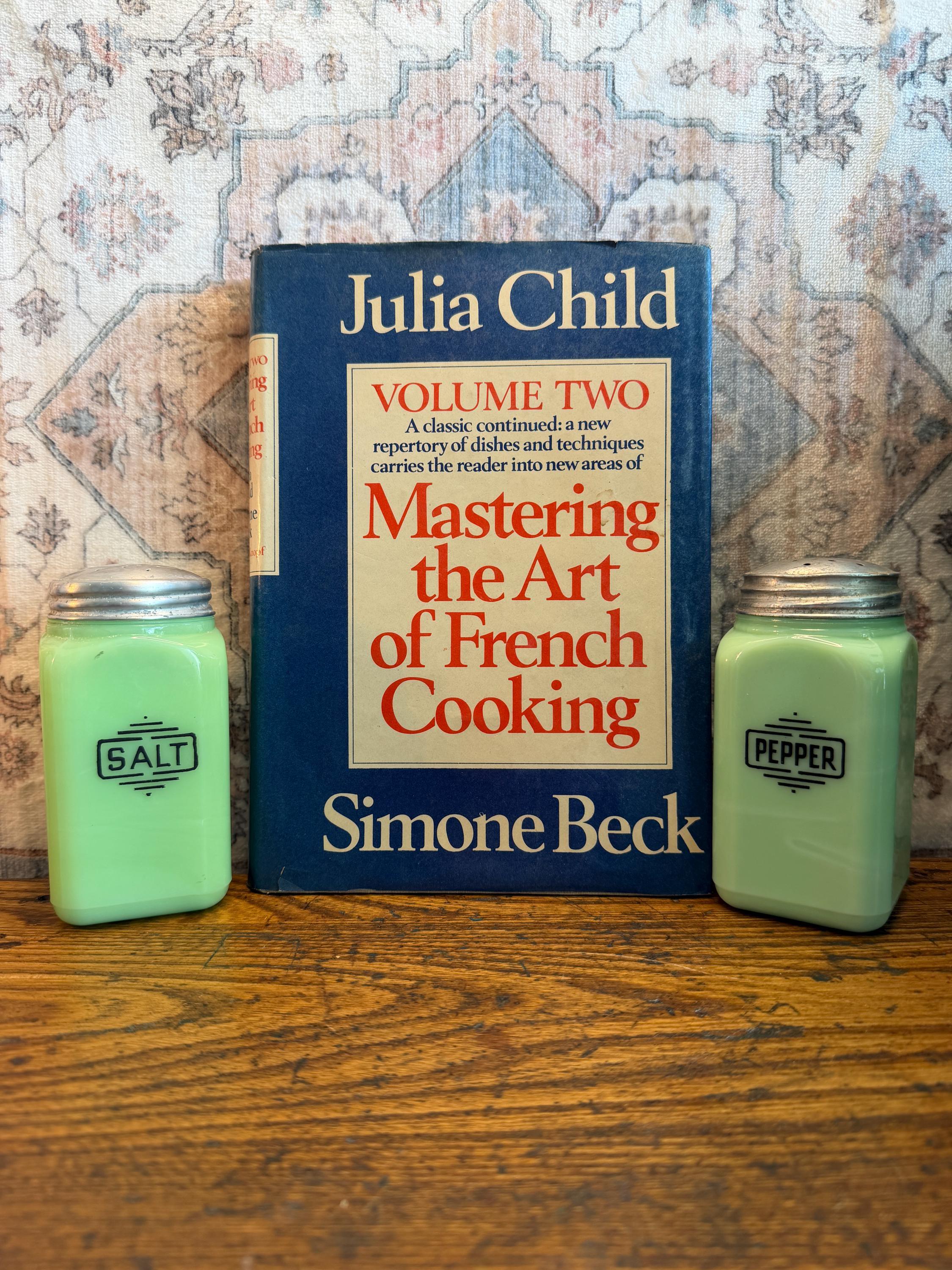 May include: A still life featuring a vintage copy of "Mastering the Art of French Cooking" by Julia Child and Simone Beck, flanked by two green glass salt and pepper shakers. The book is blue with orange text. The salt and pepper shakers have silver lids.