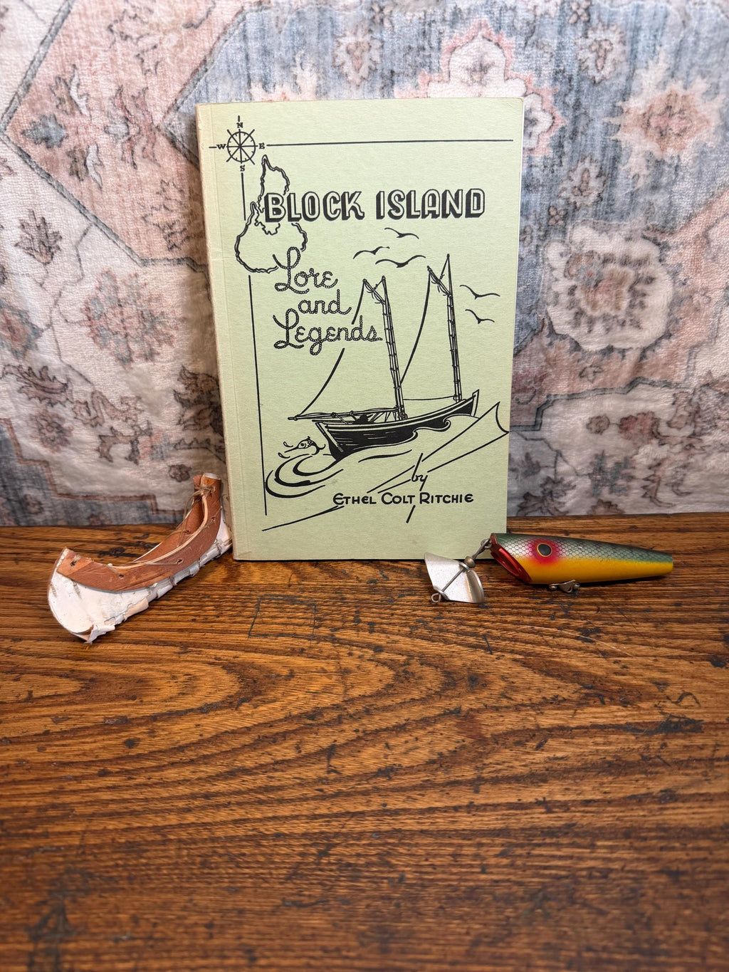 May include: A vintage book titled "Block Island Lore and Legends" with a sailboat illustration. Beside the book are a fishing lure and a small, weathered wooden boat piece, all on a wooden surface.