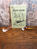 May include: A vintage book titled "Block Island Lore and Legends" with a sailboat illustration. Beside the book are a fishing lure and a small, weathered wooden boat piece, all on a wooden surface.